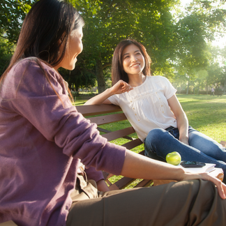 Asian women talking on bench in park with casual clothes on a sunny day, with lots of tree in background.