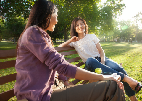 Asian women talking on bench in park with casual clothes on a sunny day, with lots of tree in background.