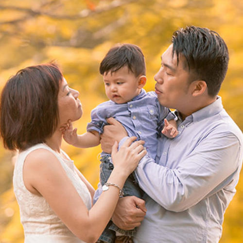 A family with father, mother, and a baby hanging around outdoors together in autumn while father is holding the baby.